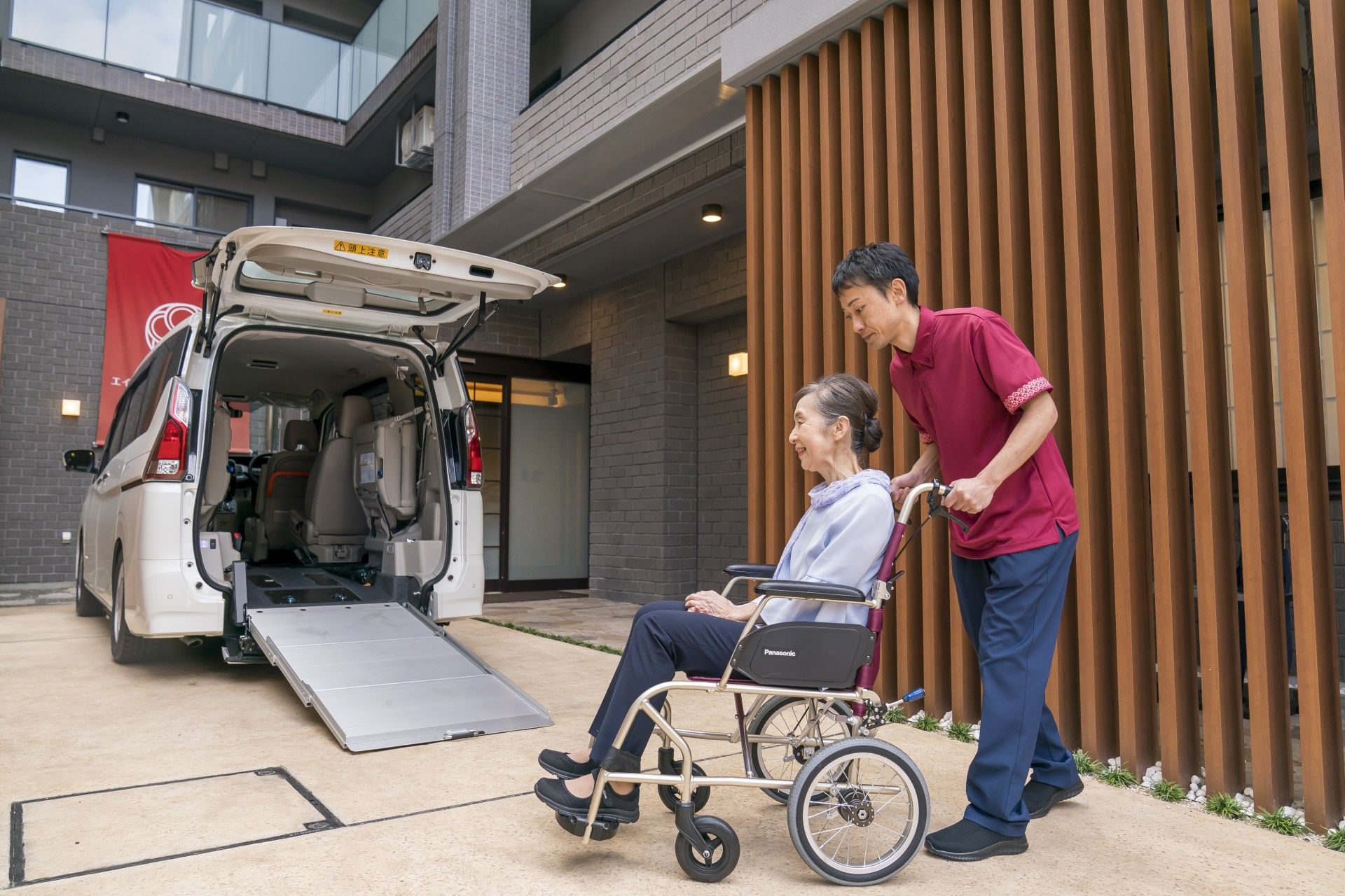 ベッドに寝ている高齢者の女性に話しかけている女性スタッフは花柄のエプロンを付けている。