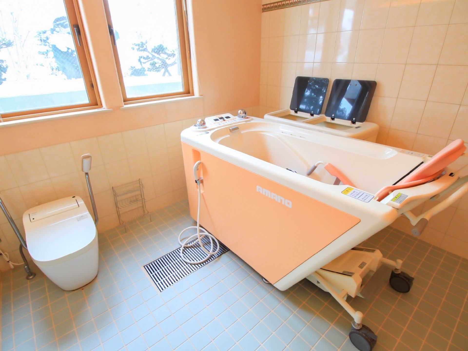 側面が薄いオレンジ色の介護浴槽がある。浴槽の隣には洋式トイレが設置され、両側に手すりが取り付けられている。