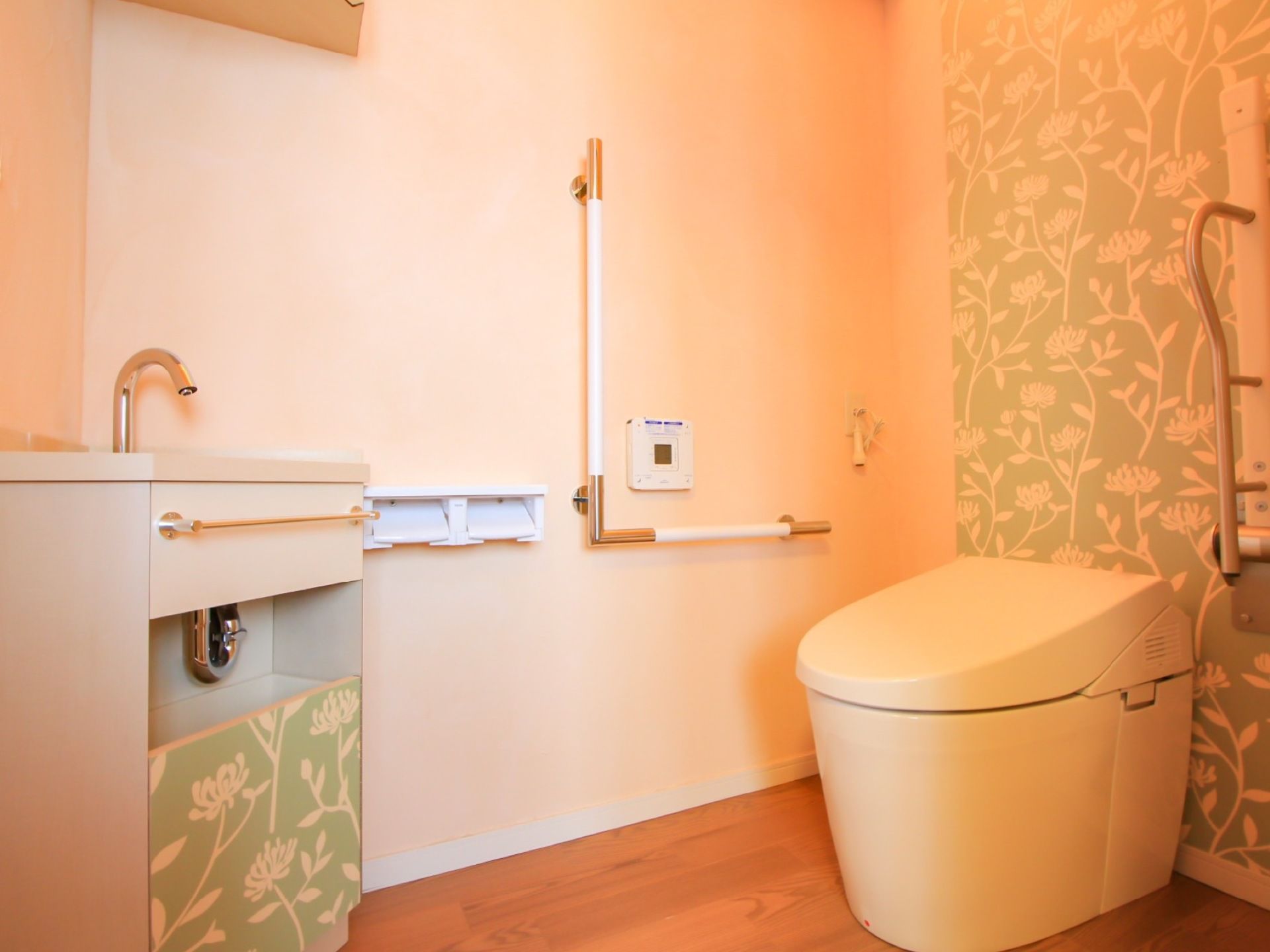 白いタンクレスのトイレがあり、花柄の壁紙が貼られている。Ｌ字型に手すりが設置され、２連タイプのペーパーホルダーが取り付けられている。