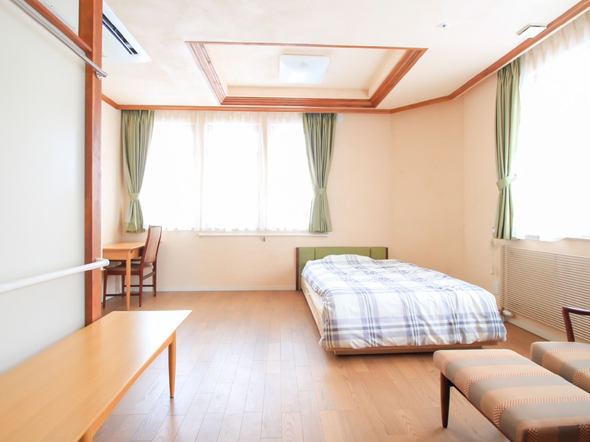 レースカーテンが掛けられた腰高窓が部屋の２面に設置されている。チェックの布団が掛けられたベッドと机や椅子が置いてある。