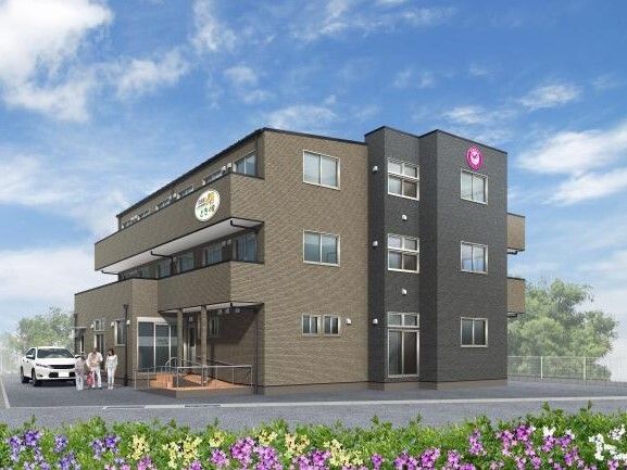 周辺には緑が多く、花が植えられているブラウン系の3階建ての建物。各フロアにベランダがあり、敷地内には車が止まっている。