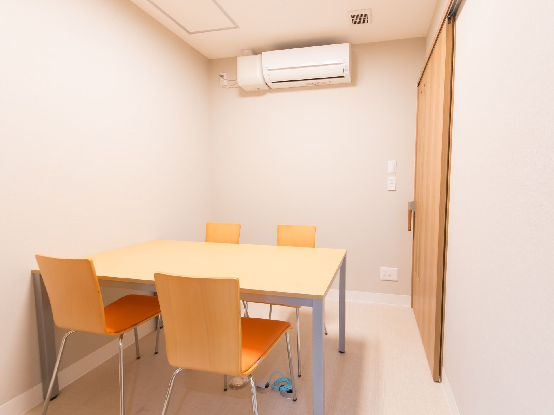 白でまとめられた内装の部屋に、木調のテーブルセットが置いてある。エアコンと換気システムが設置されている。