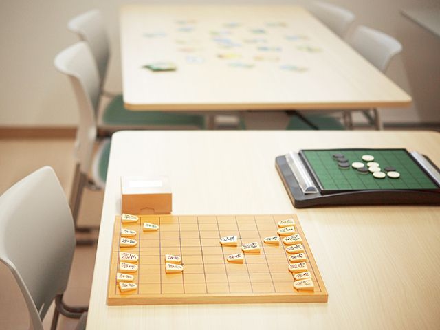 4脚ずつ椅子がセットされオセロや将棋が並べられたテーブルの後ろにあるテーブルには、何かのカードゲームのカードが並べられている。