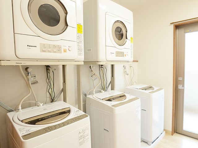 乾燥機が二台、縦型の洗濯機が三台設置された洗濯室。壁は白く、床は明るい色のフローリングで余計なものは置かれていない。