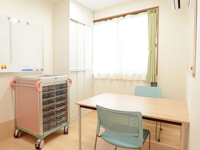 たくさんの引き出しが付いたワゴンが置かれ、その横には向かい合わせに2脚の椅子がセットされたテーブルが置かれ、壁にはホワイトボードの予定表がある。