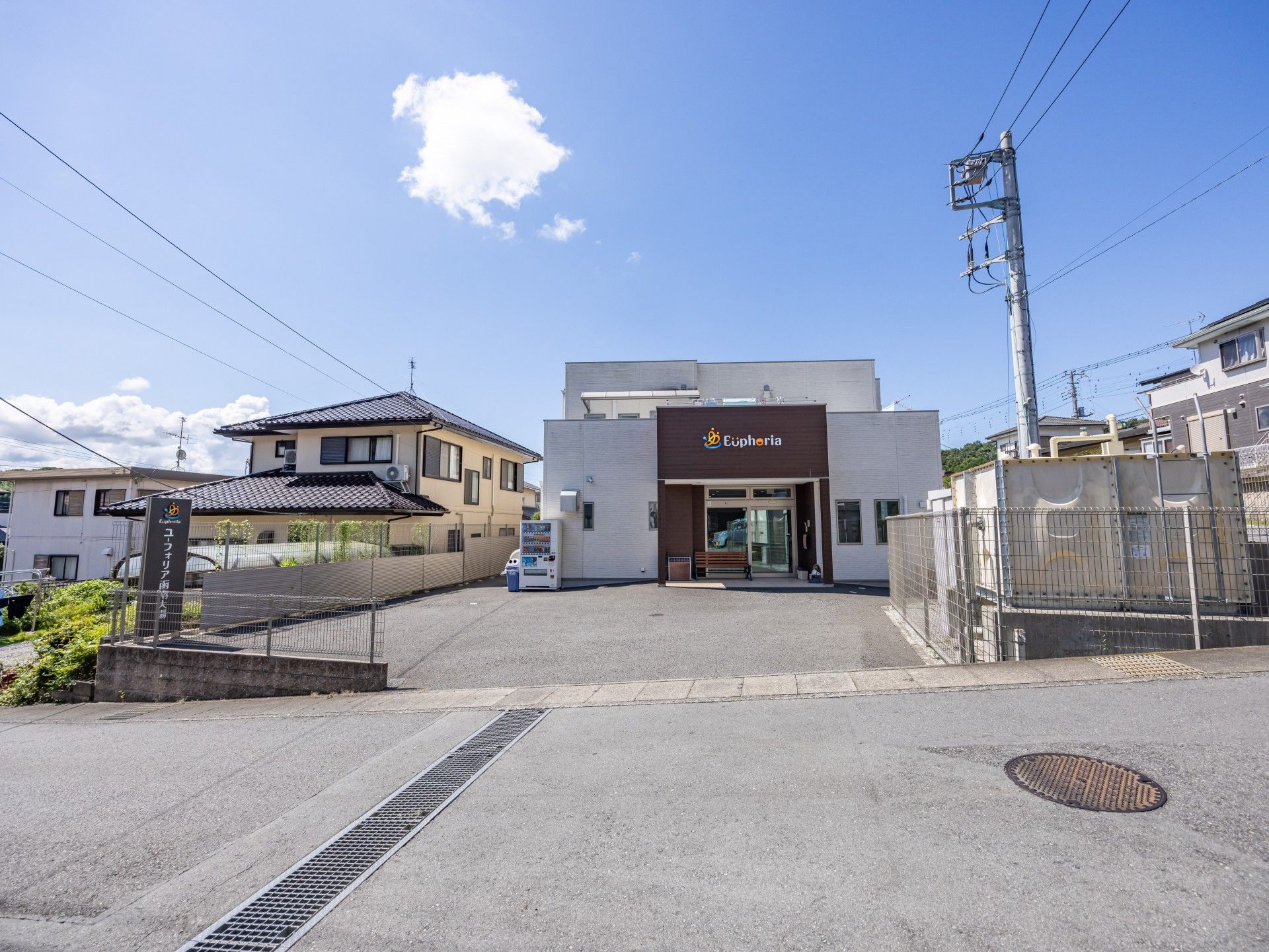 白とブラウンの建物。広い駐車場があり、自動販売機が玄関のそばにあり、出入り口の横には木製のベンチも置かれ、車寄せが付いている。