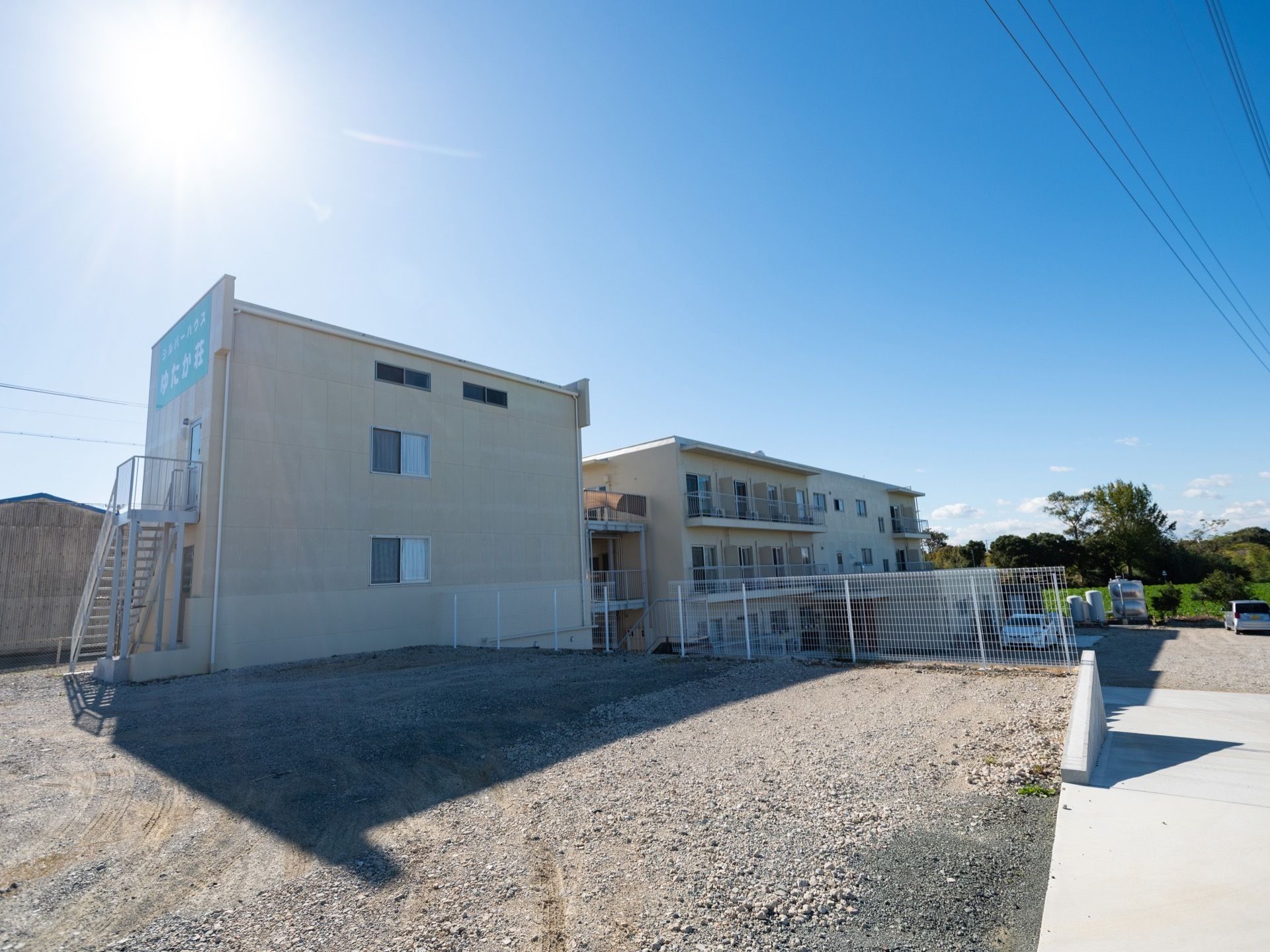 白い壁の２棟の建物が建っている。手前の建物には、外階段が設置されている。階段の上の壁に大きく施設名が入っている。