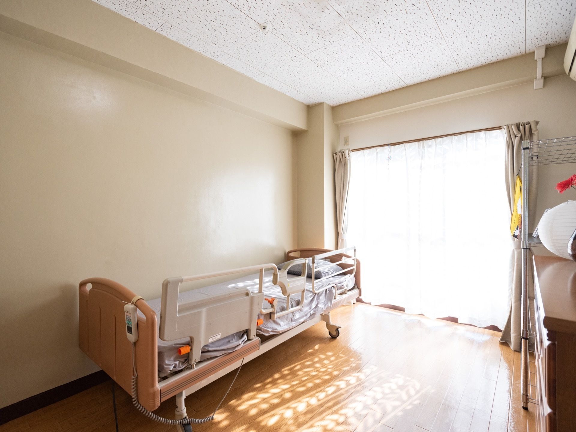 広い窓から陽光が差し込む明るい部屋である。ベッドは、リモコンで操作可能な電動式介護ベッドとなっている。転落防止の柵が付いている。