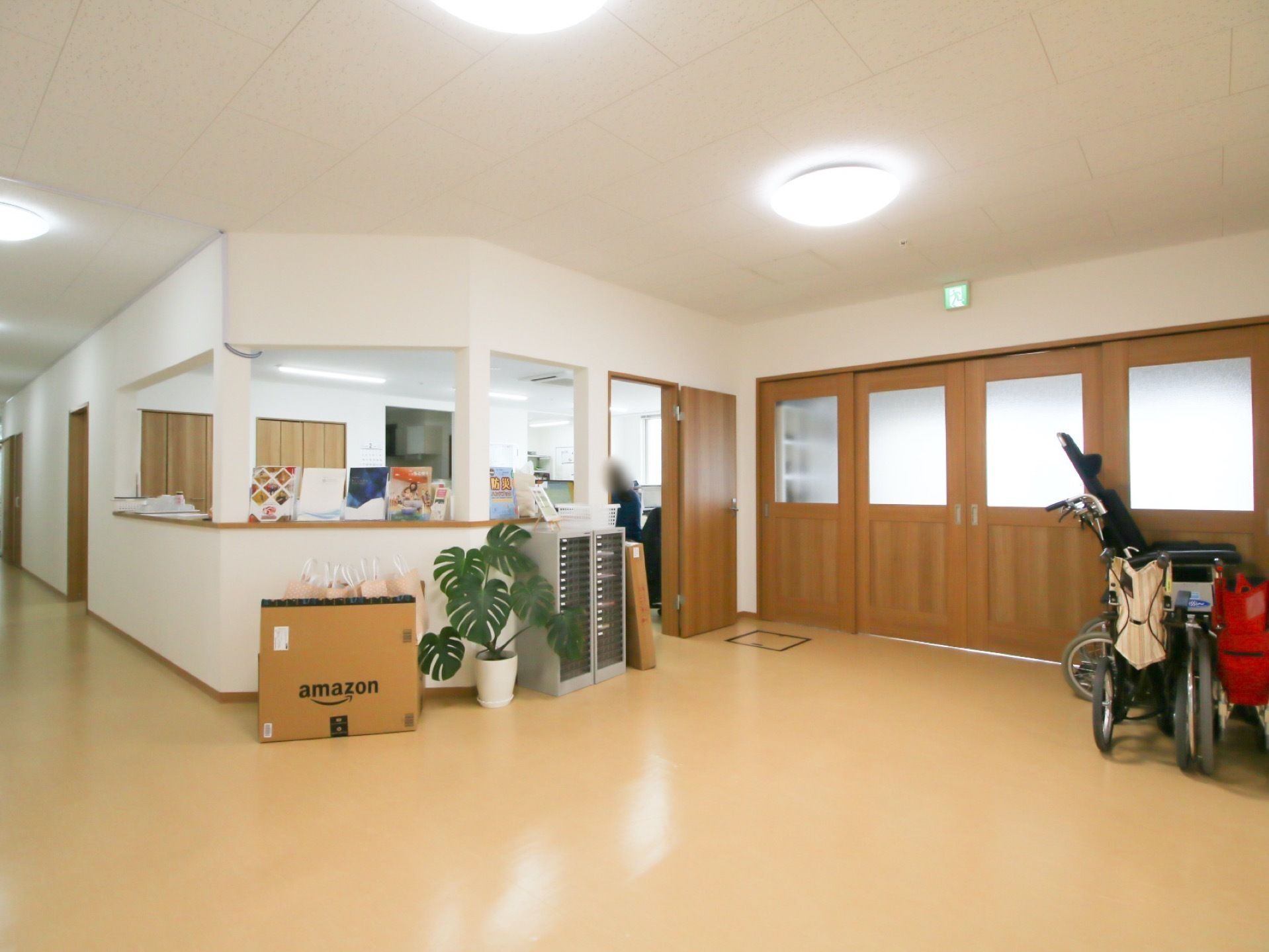 風除室からすぐの所に事務室がある。通路側のカウンター上に書類が置かれている。部屋の中から通路を往来する入居者様や来訪者様の確認が可能である。