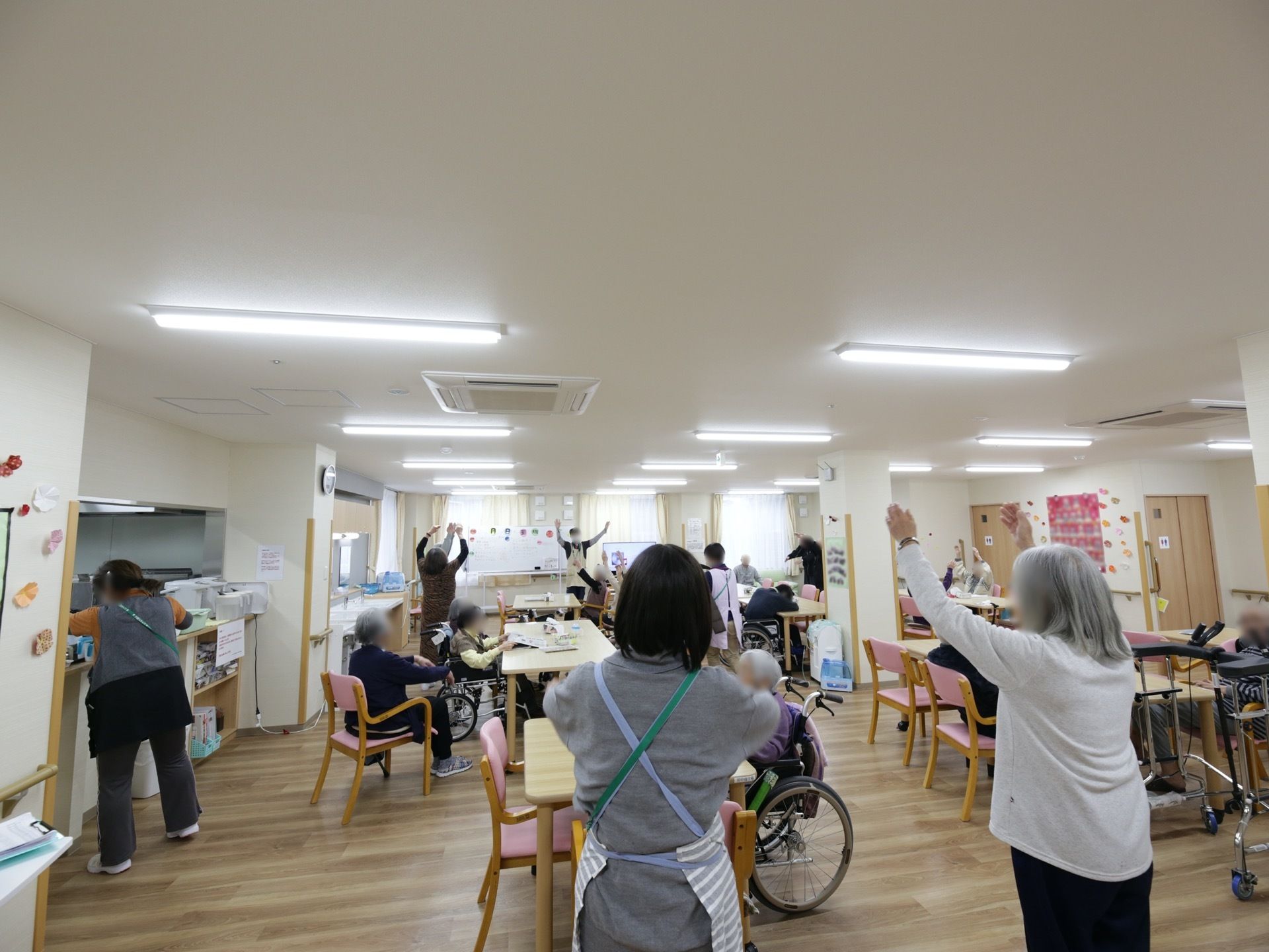 共有スペースで入居者様とスタッフが集い、歓談したり、体操したりしている。ダイニングテーブルが間隔を空けて配置されている。