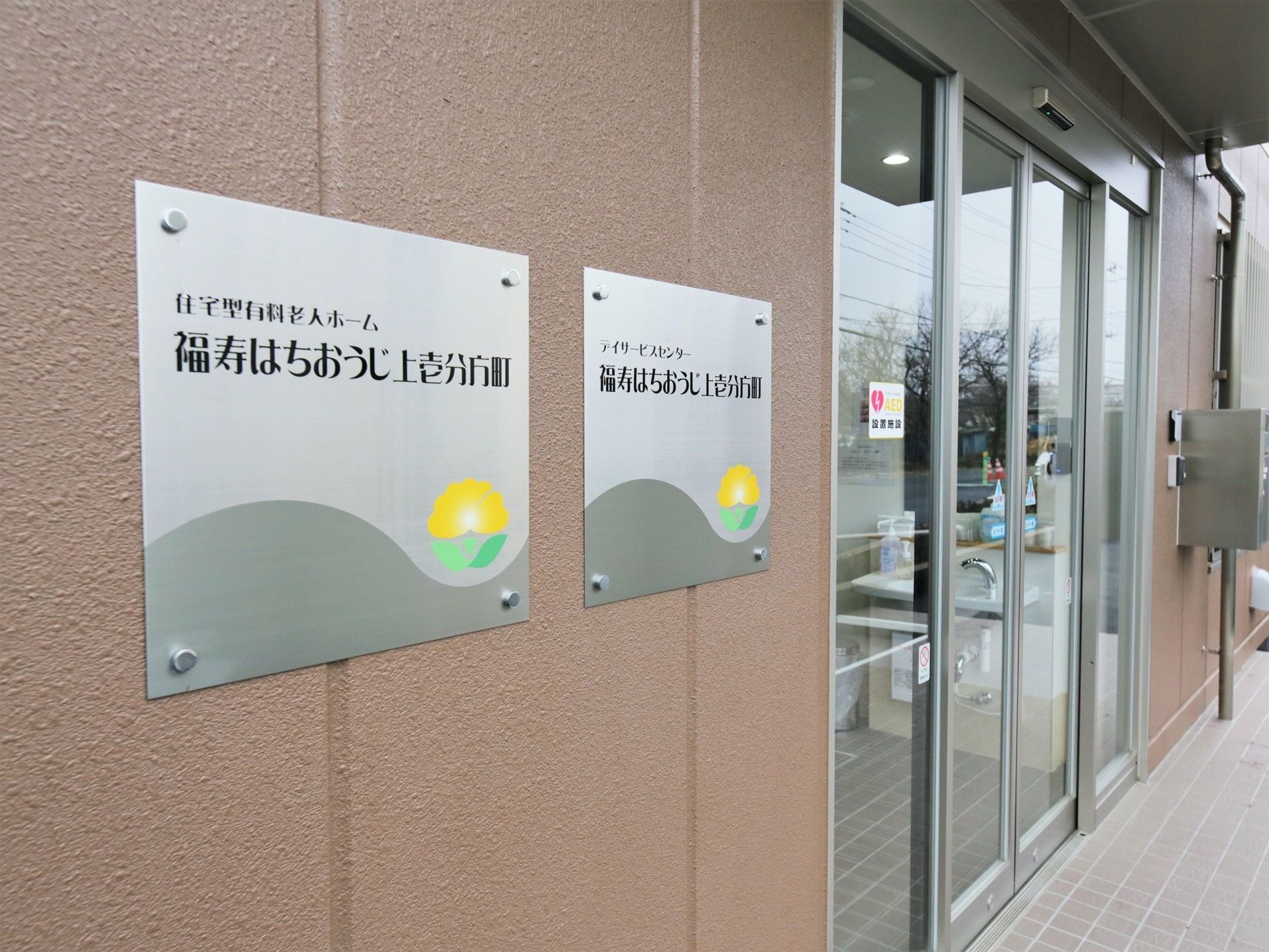 玄関脇の壁に施設名が表示されている。ステンレス製のプレートが掲げられている扉は、両開きの自動ドアになっている。