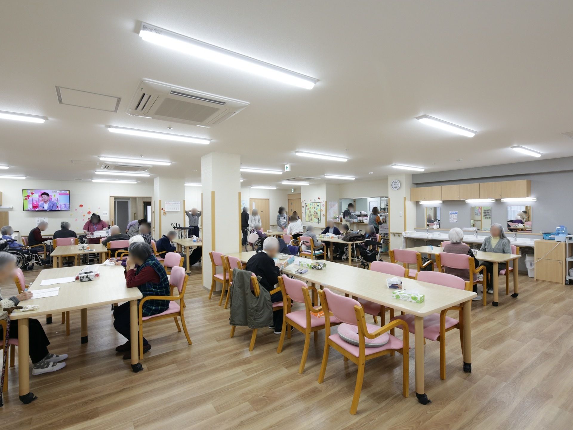 入居者様たちが、共有スペースに置かれているダイニングテーブルを囲んで寛いでいる。コーナーには洗面台が設置されている。