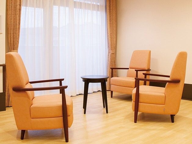オレンジの肘置き付きの大きな一人用の椅子がテーブルを囲むように3脚設置されている。大きな窓が付きレースカーテンとオレンジのカーテンが付いている。