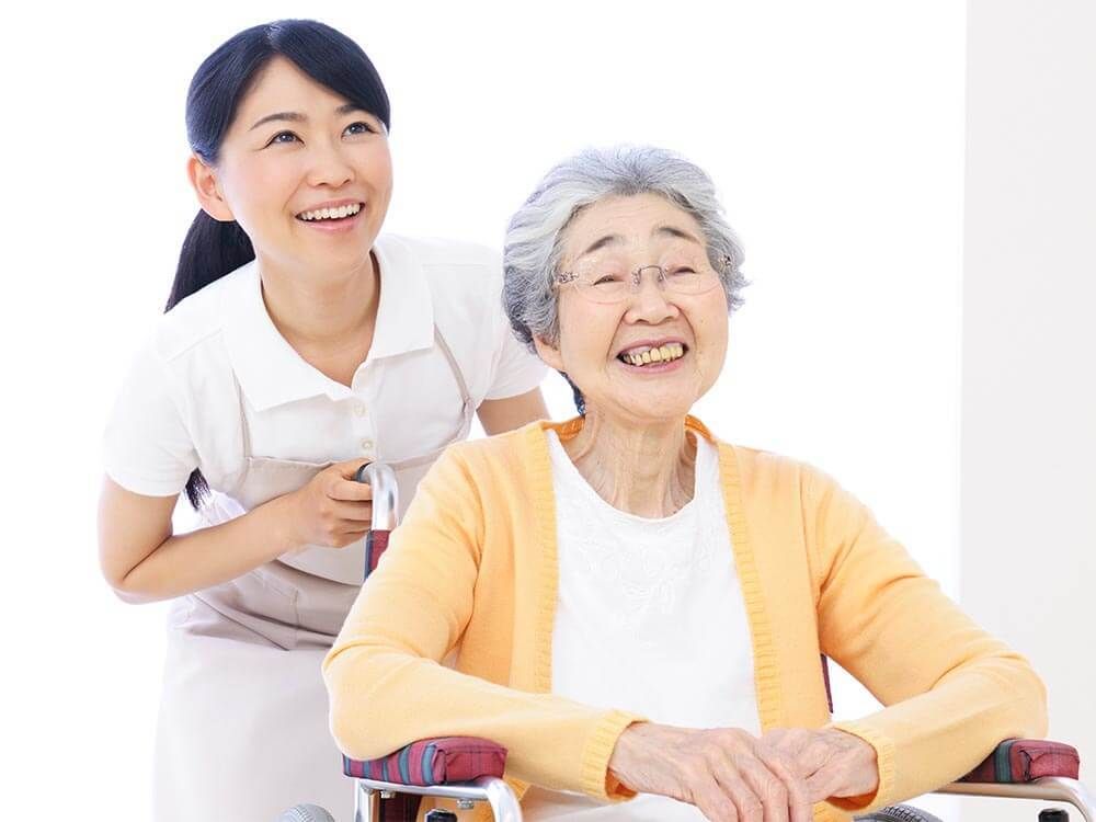 笑顔で車いすに座っている眼鏡をかけた高齢女性と車いすを押している白衣を着た女性スタッフ。