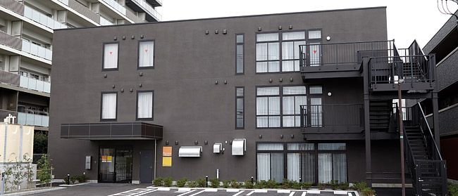 白衣を着た女性スタッフが車いすを押し、カーディガンを着た高齢女性が車いすに座っている。