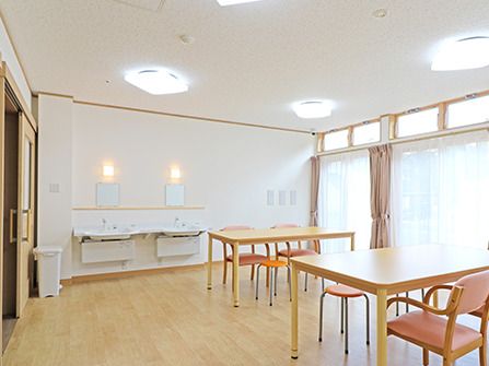 レースカーテンとカーテンが付いた大きな窓がある食堂兼談話室には車いす対応の洗面所があり、ダイニングテーブルが複数設置されている。
