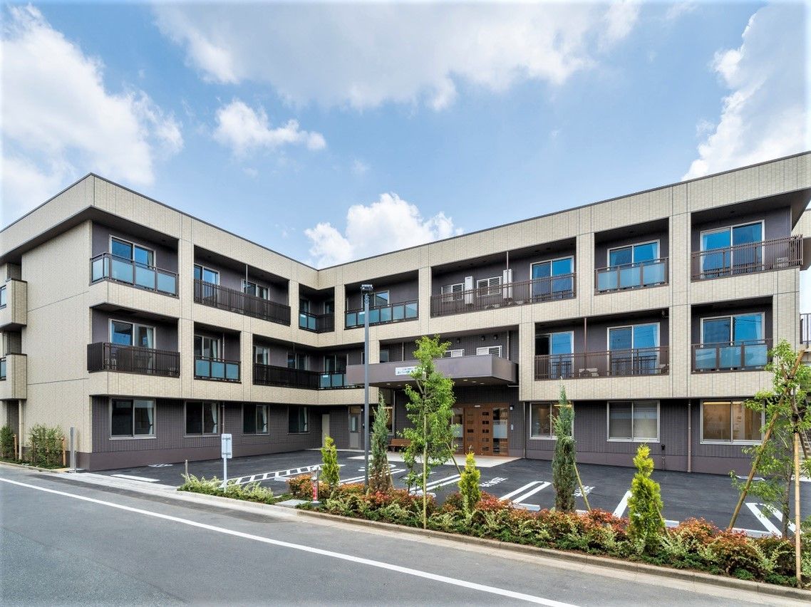 3階建ての建物はエル字型でベランダが付き、花壇で囲まれた駐車場もある。