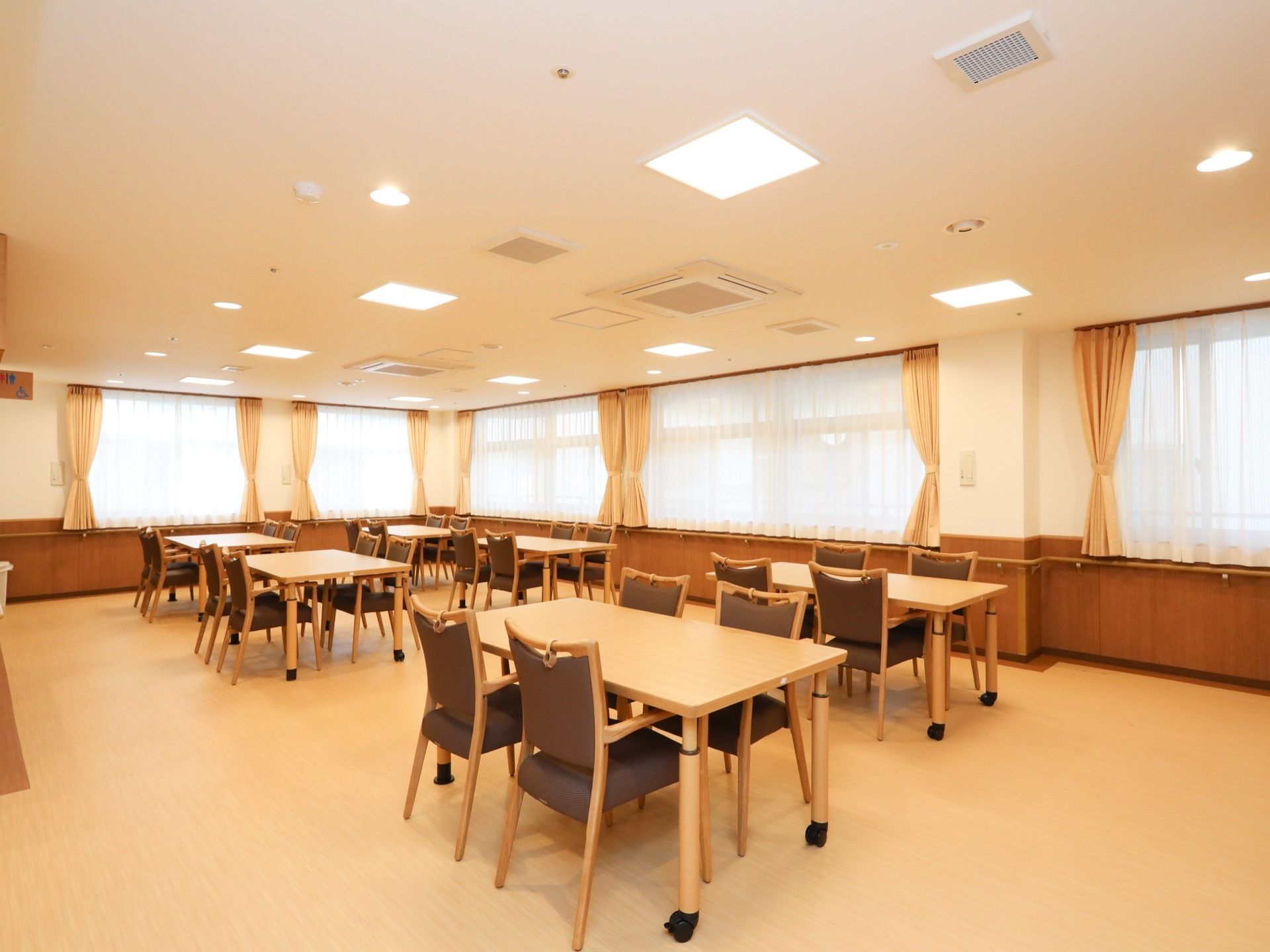 明るく広々とした食堂には、キャスター付きの４人掛けテーブルが並べられている。手すりが壁に沿って取り付けられている。