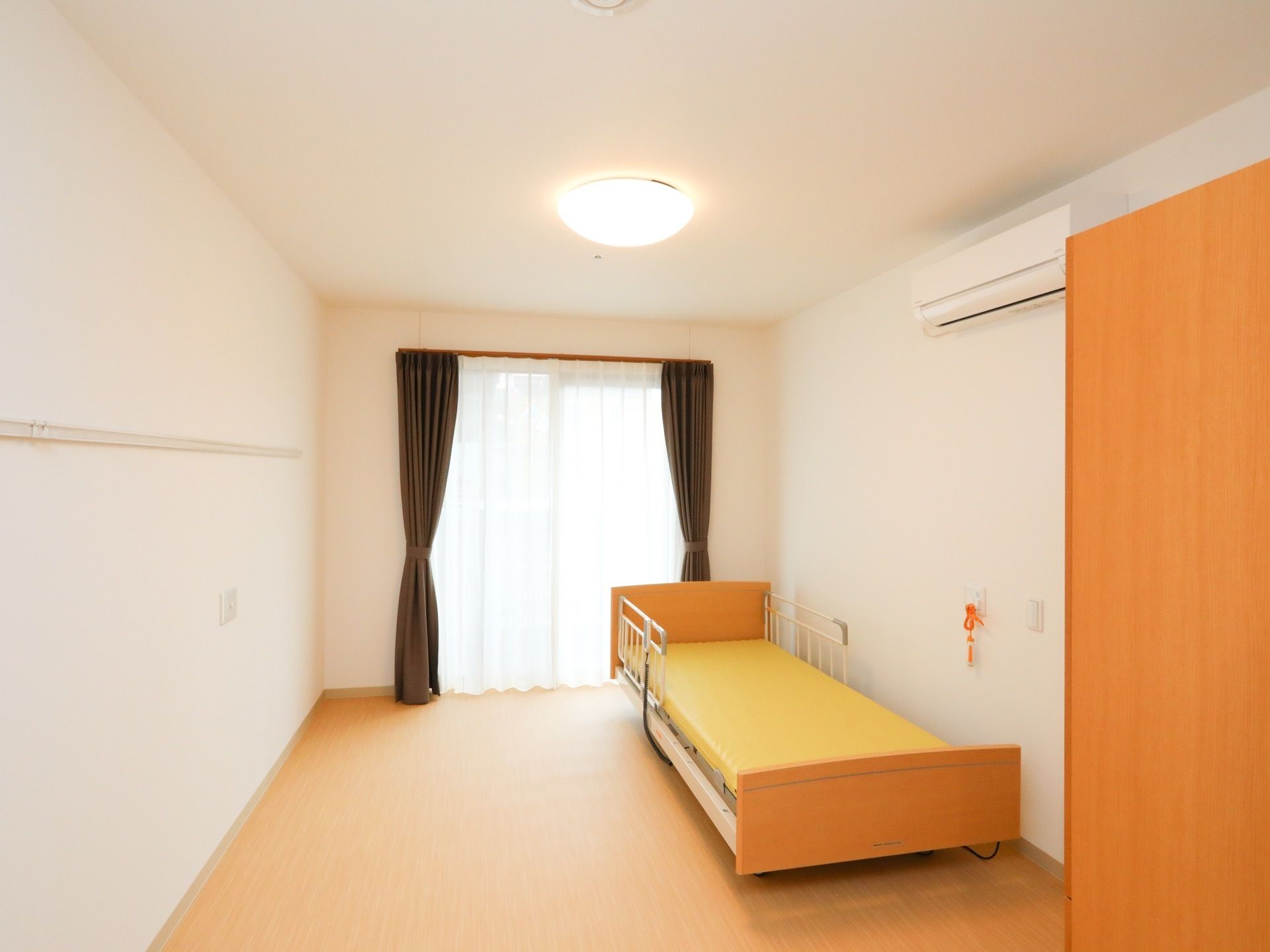 掃き出し窓が設けられた明るい居室である。電動介護ベッドやナースコール、エアコンが完備されており、収納家具も用意されている。