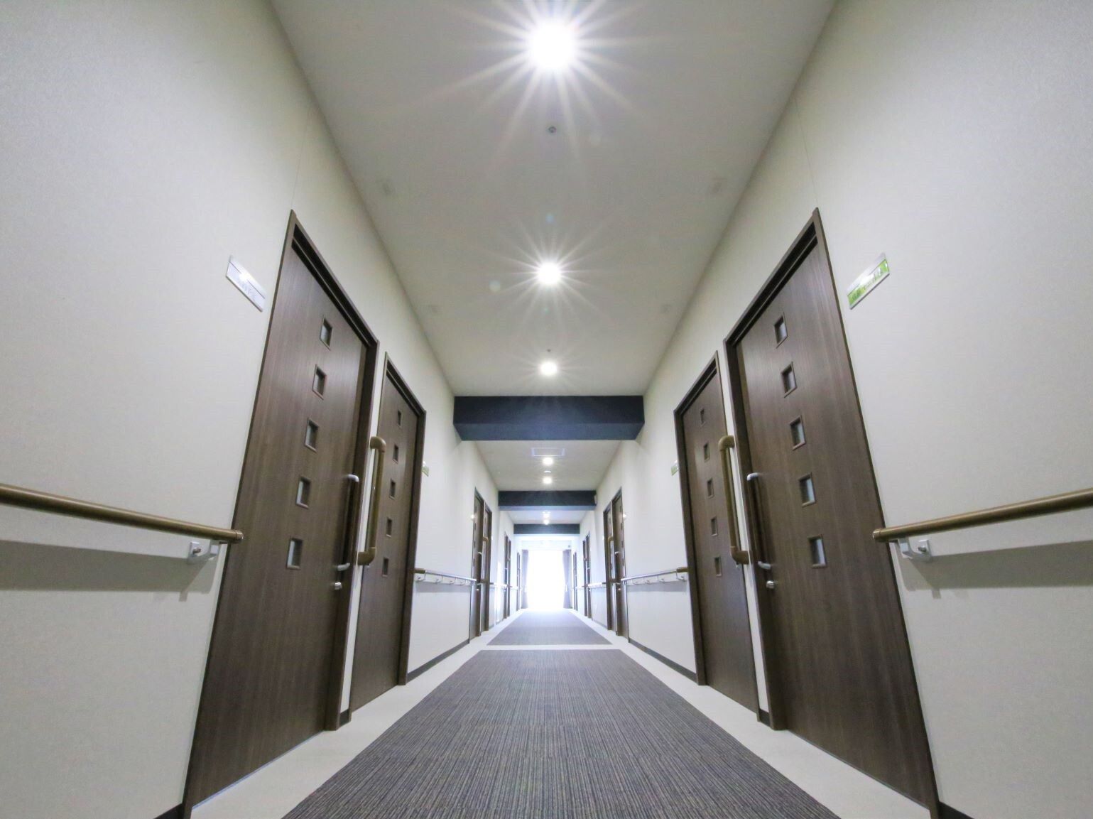 まっすぐな廊下の両サイドに手すりと居室のドアがあり、突き当たりの大きな窓から陽の光が差し込んでいる。居室のドアの横には小さなプレートがある。