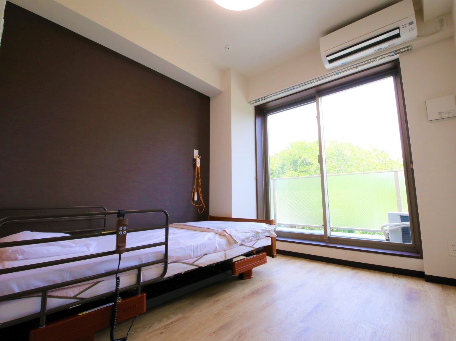 エアコンが付いたシンプルなフローリングの部屋には寝具付きのベッドが置かれ、壁には緊急通報装置が付き、大きな窓も付いている。