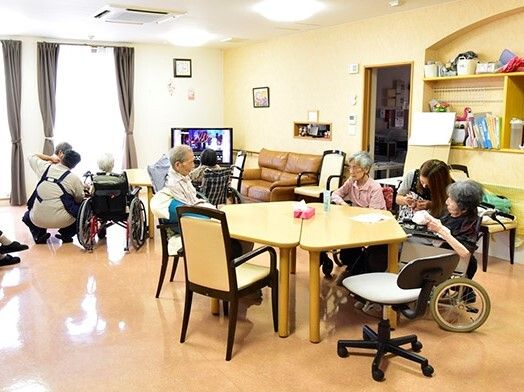 スタッフと入居者が笑顔で過ごしているリビングダイニング。テレビやソファもあり、テーブルの上にはティッシュがある。