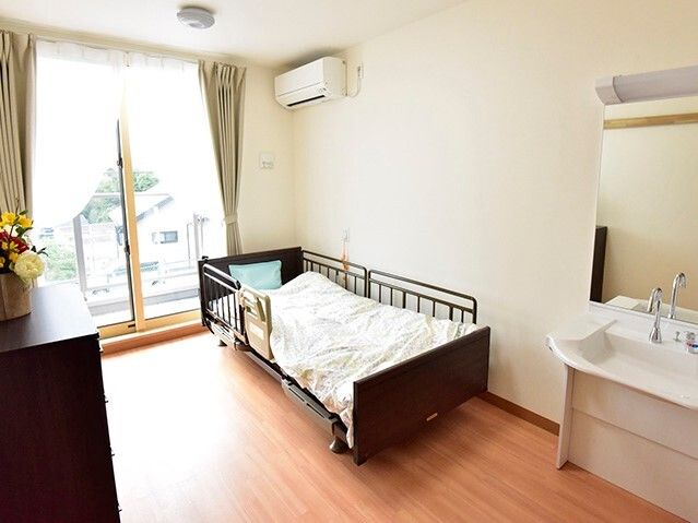 寝具付きのベッドや車いす対応の洗面所、タンスなどが置かれたフローリングの部屋には大きな窓があり、カーテンも付いている。