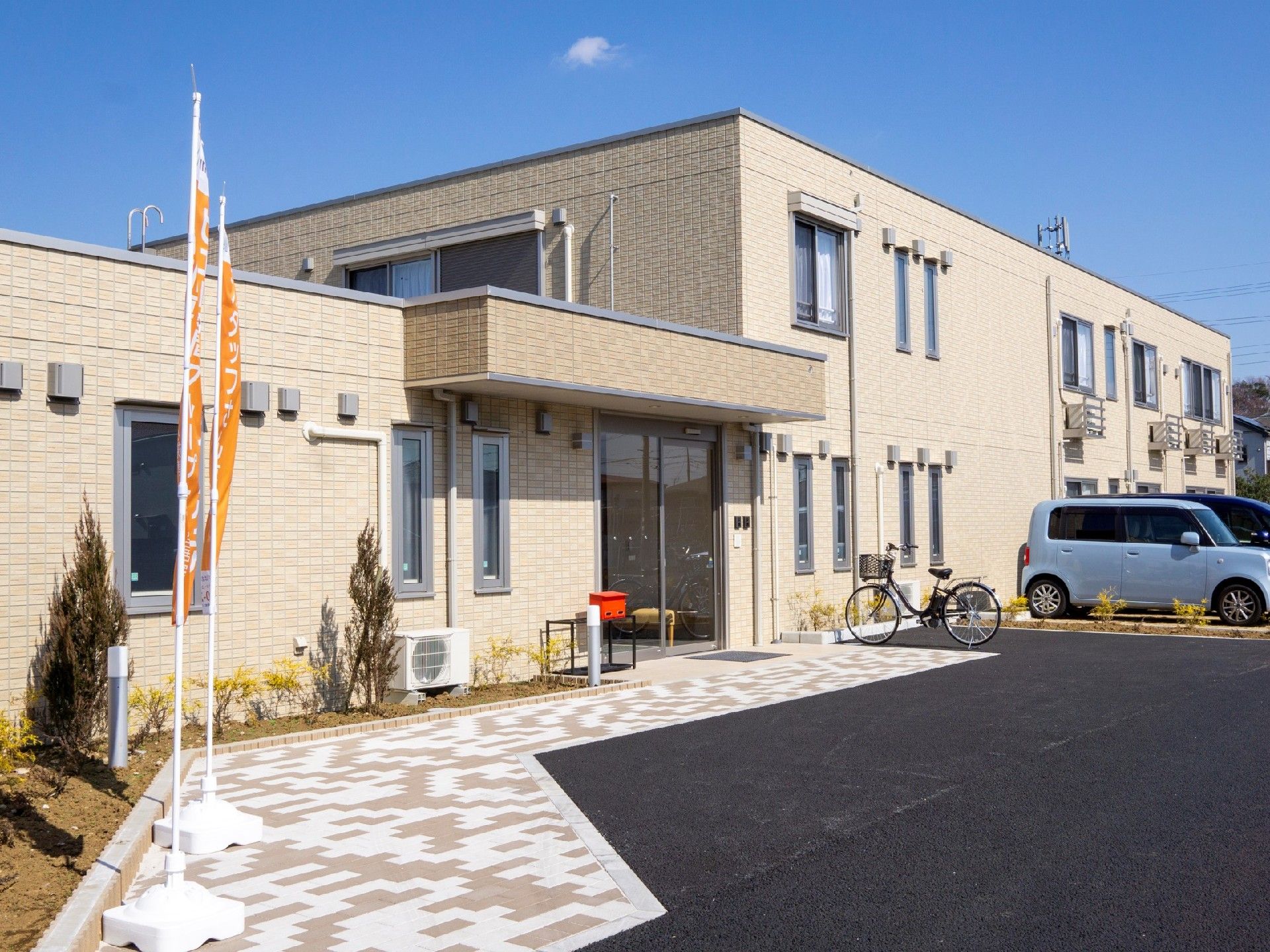ベージュですっきりとした建物は一部2階建てで、周囲には木が植えられ、駐車スペースもある。