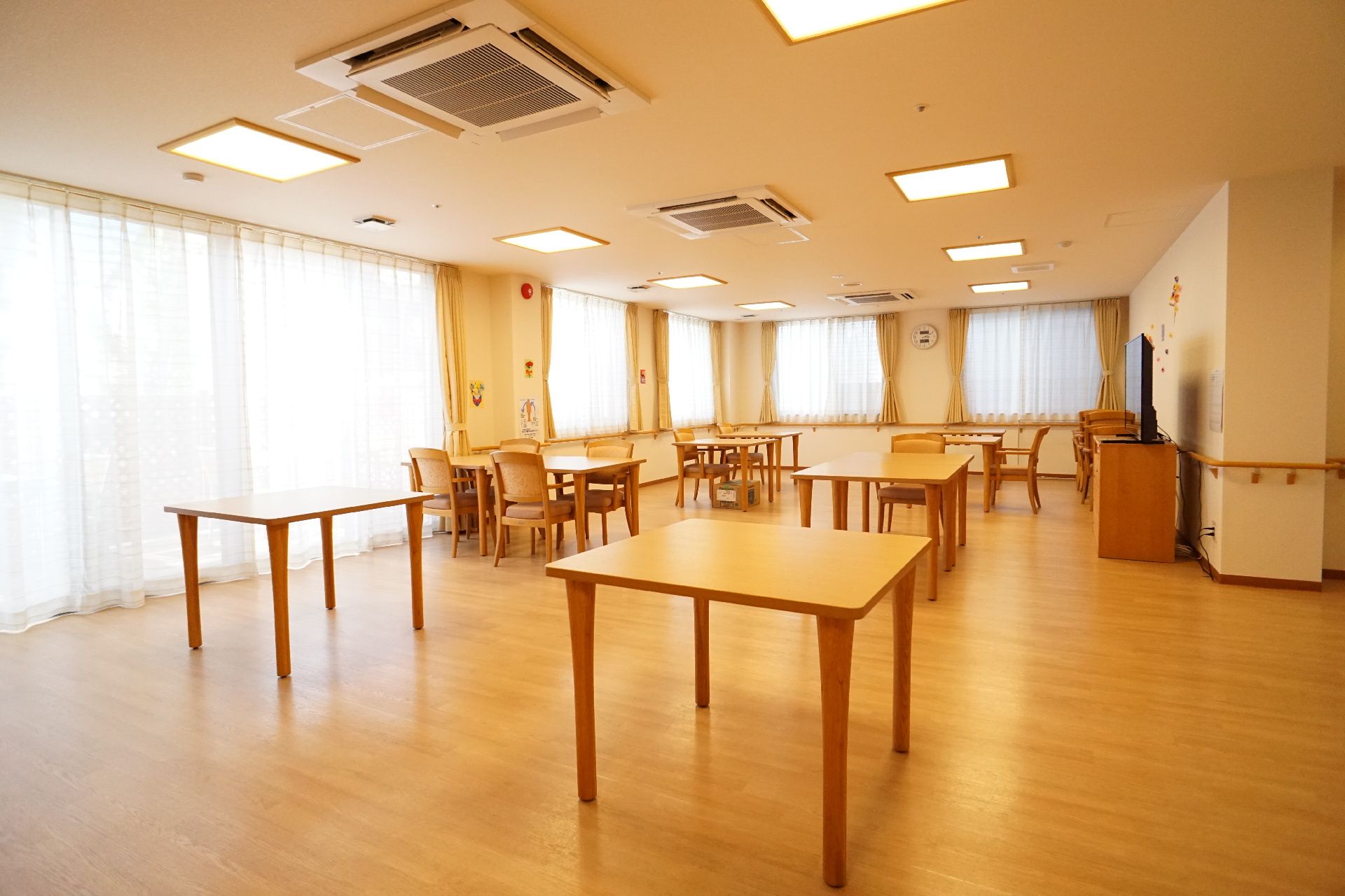 窓に囲まれた部屋に並んでいる木目調の机と椅子