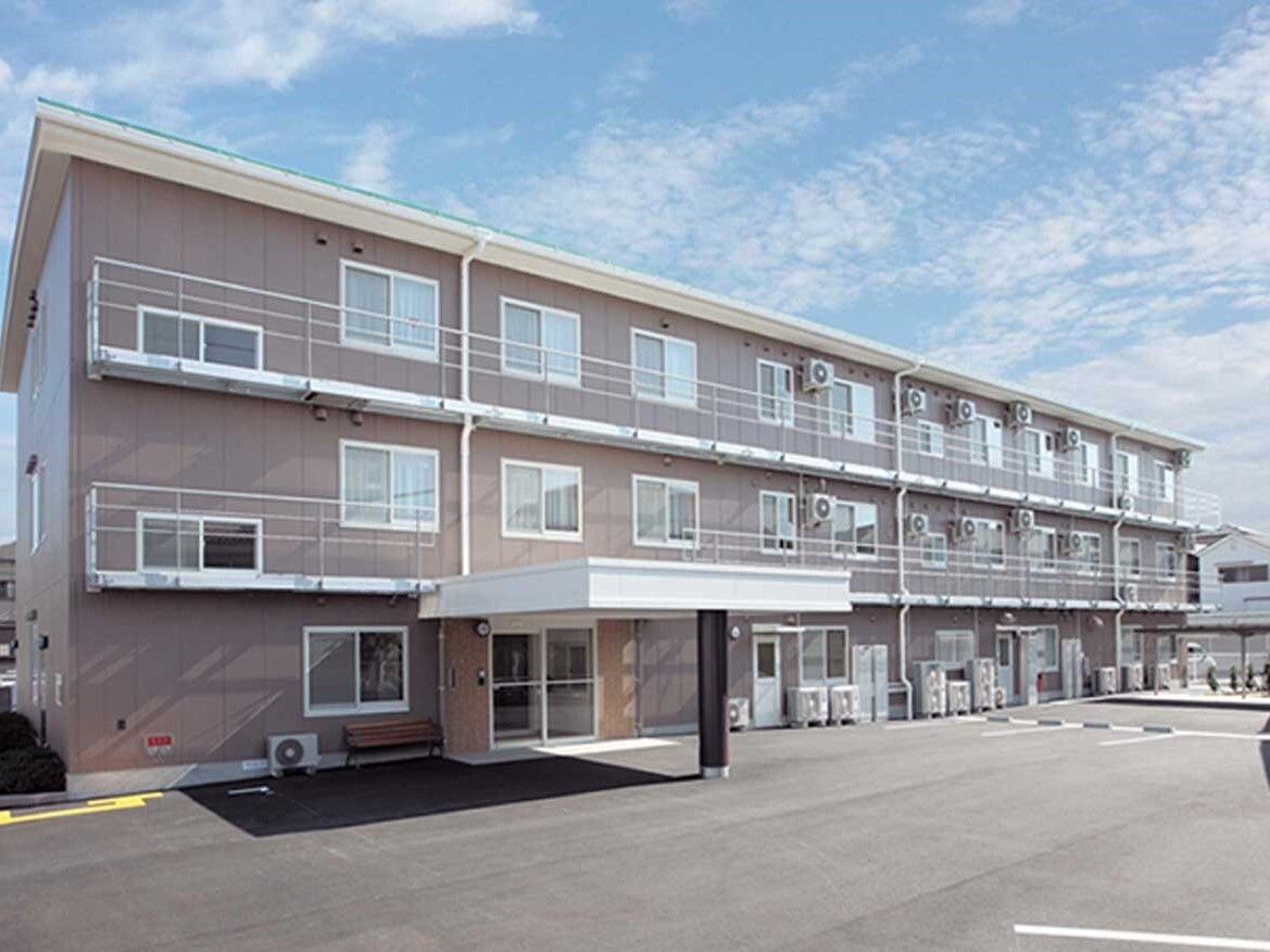 グレー系の３階建ての建物。玄関先には車寄せがあり、広い駐車場が設置され、各フロアにベランダが付いている。