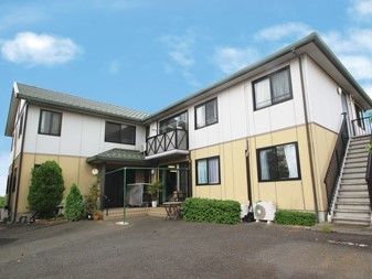 1階部分がベージュ、2階部分が白い2階建ての建物。玄関には屋根が付き、2階部分には一部ベランダがある。