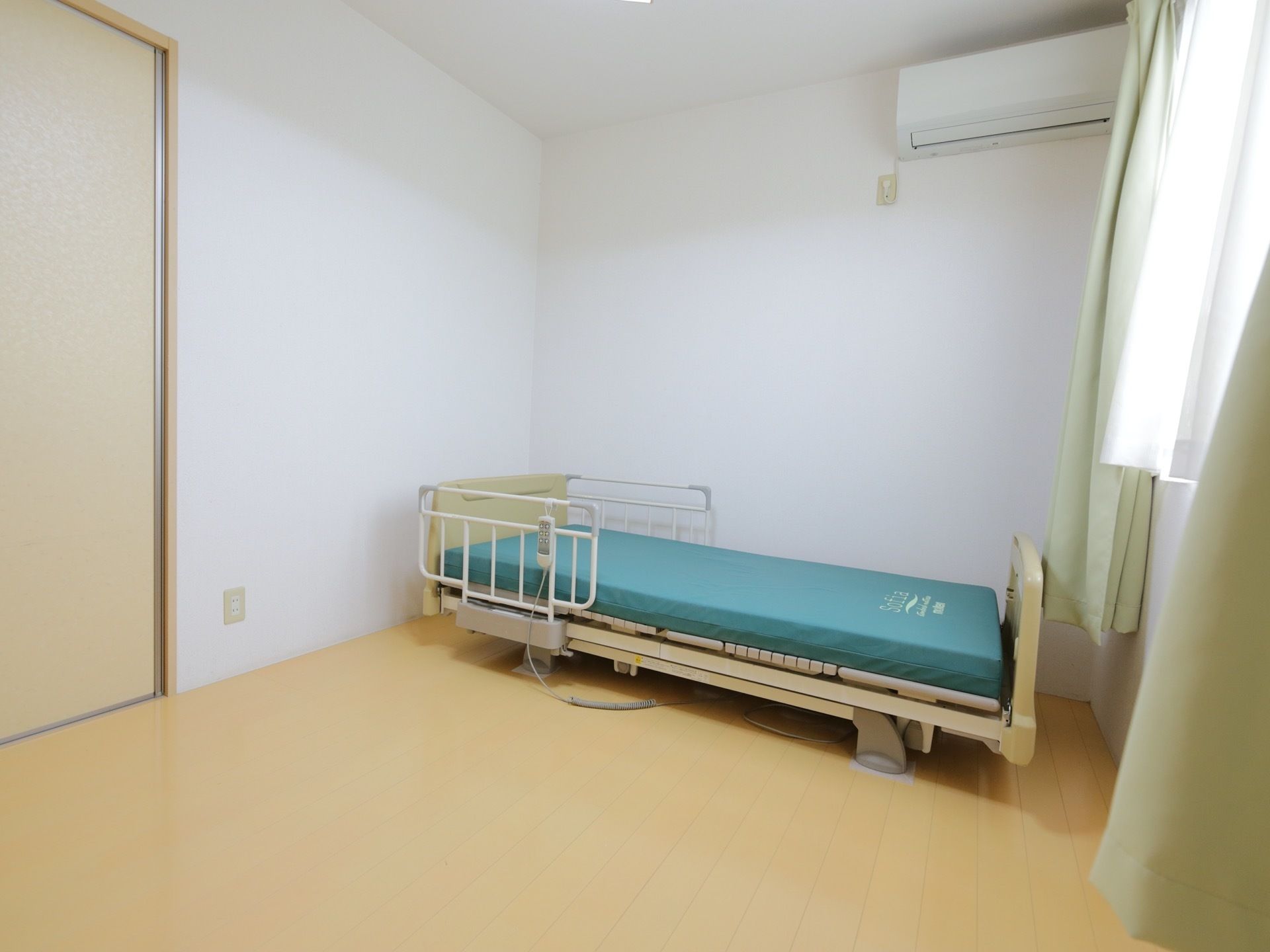 明るい色調の居室である。転落防止柵がついた介護ベッドが壁際に置かれている。操作用リモコンが柵に掛けられている。