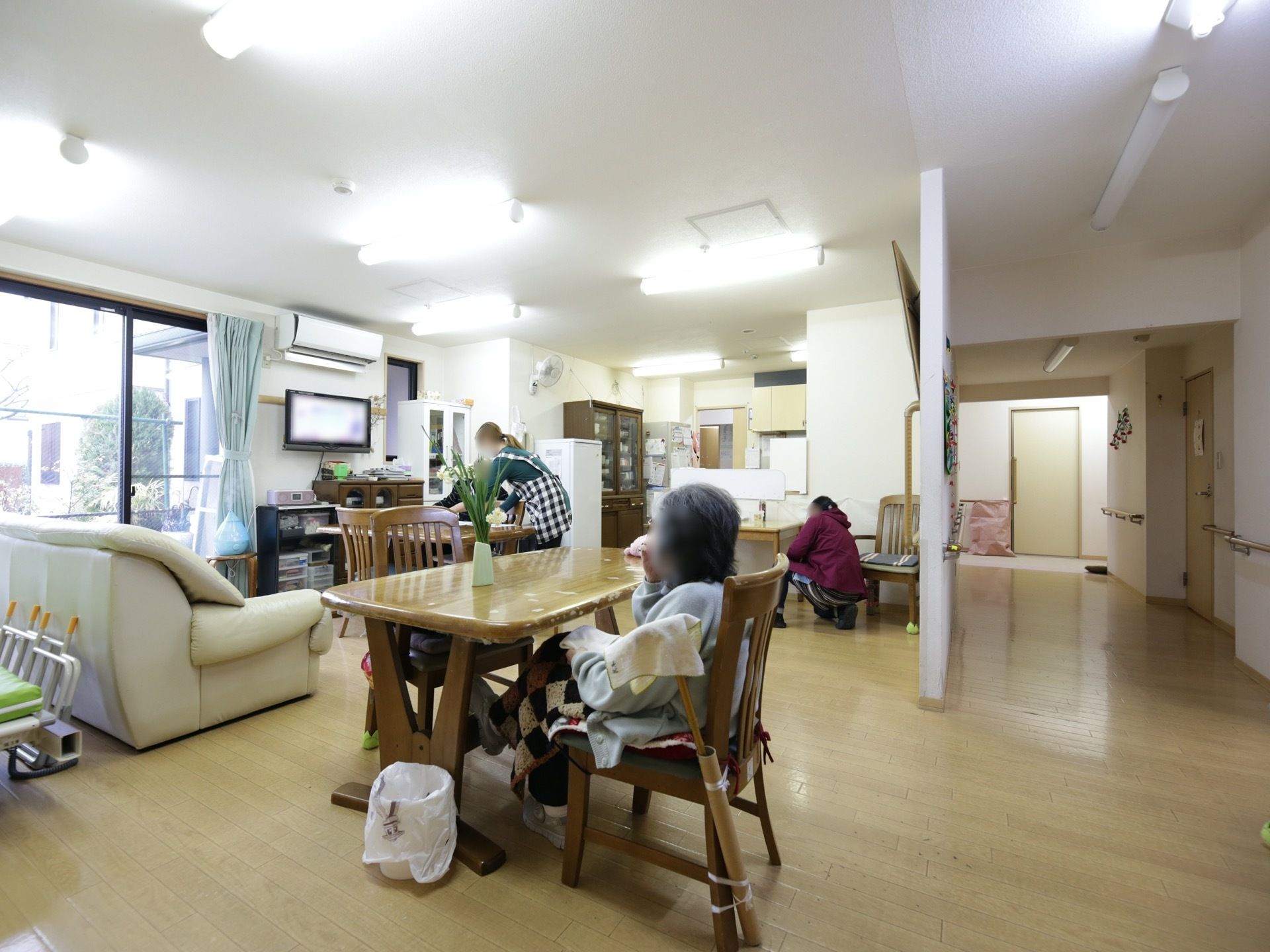食堂のテーブルにある椅子に女性の入居者様が１名座っている。奥の方で、２名の女性スタッフが作業をしている。