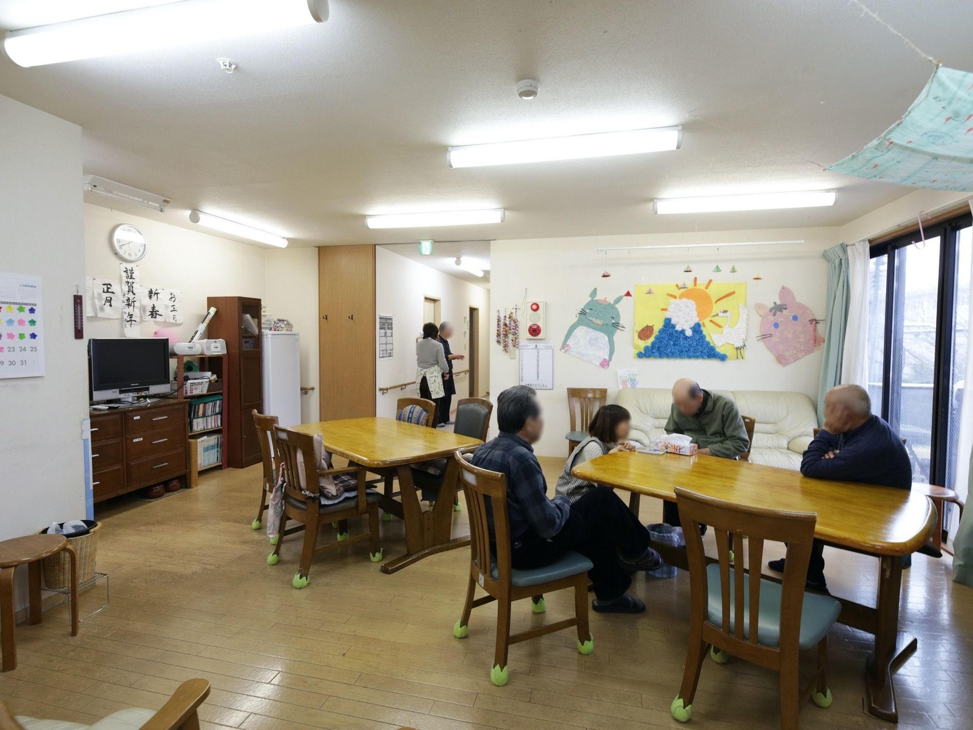 食堂の窓際にあるテーブルに男性の入居者様が３名集まっている。廊下では、スタッフが入居者様と立ち話をしている。