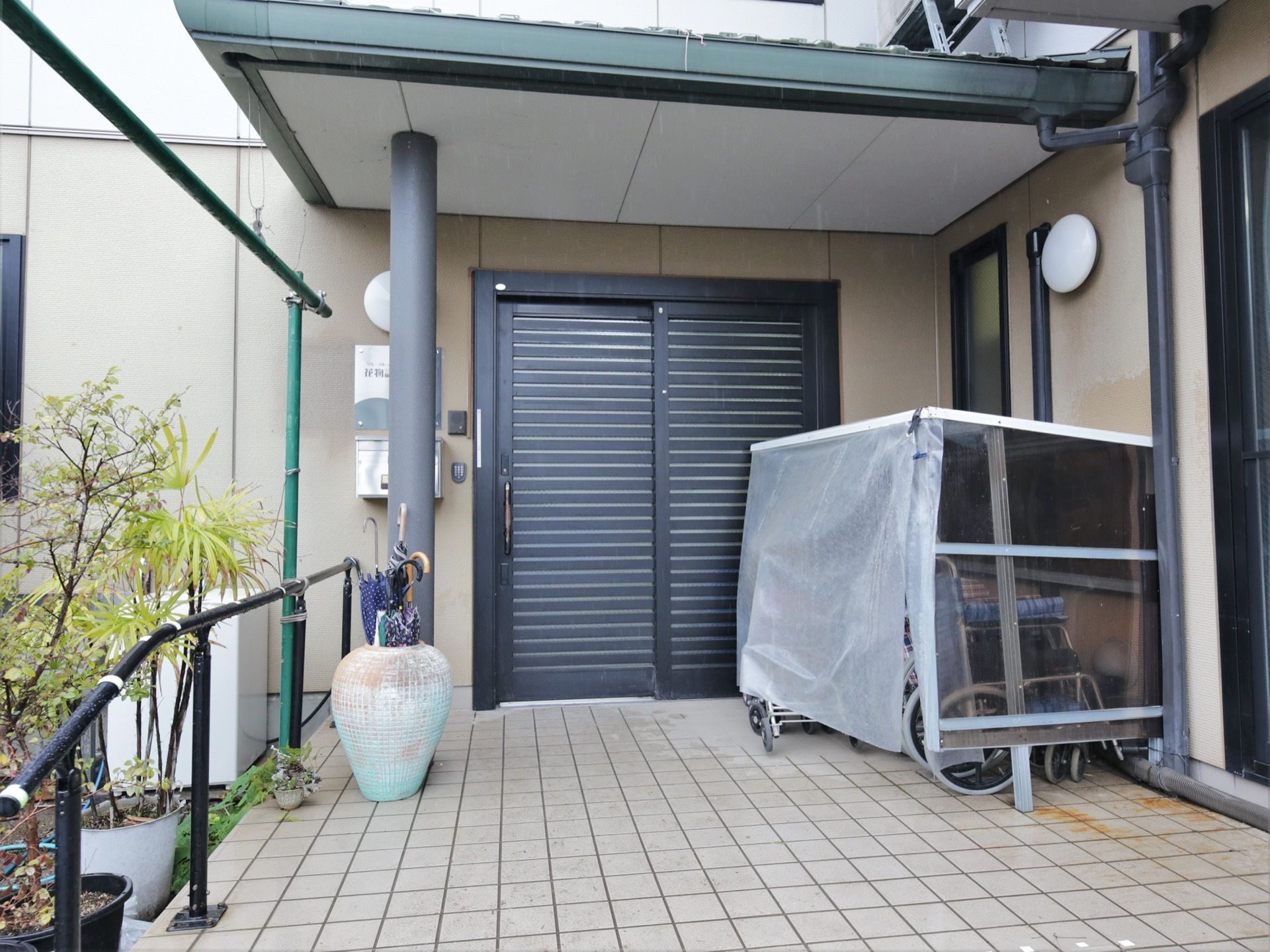 玄関は横方向に枠がついた引き戸である。玄関前のひさしの下にある物置の中に車椅子が何台か置かれている。