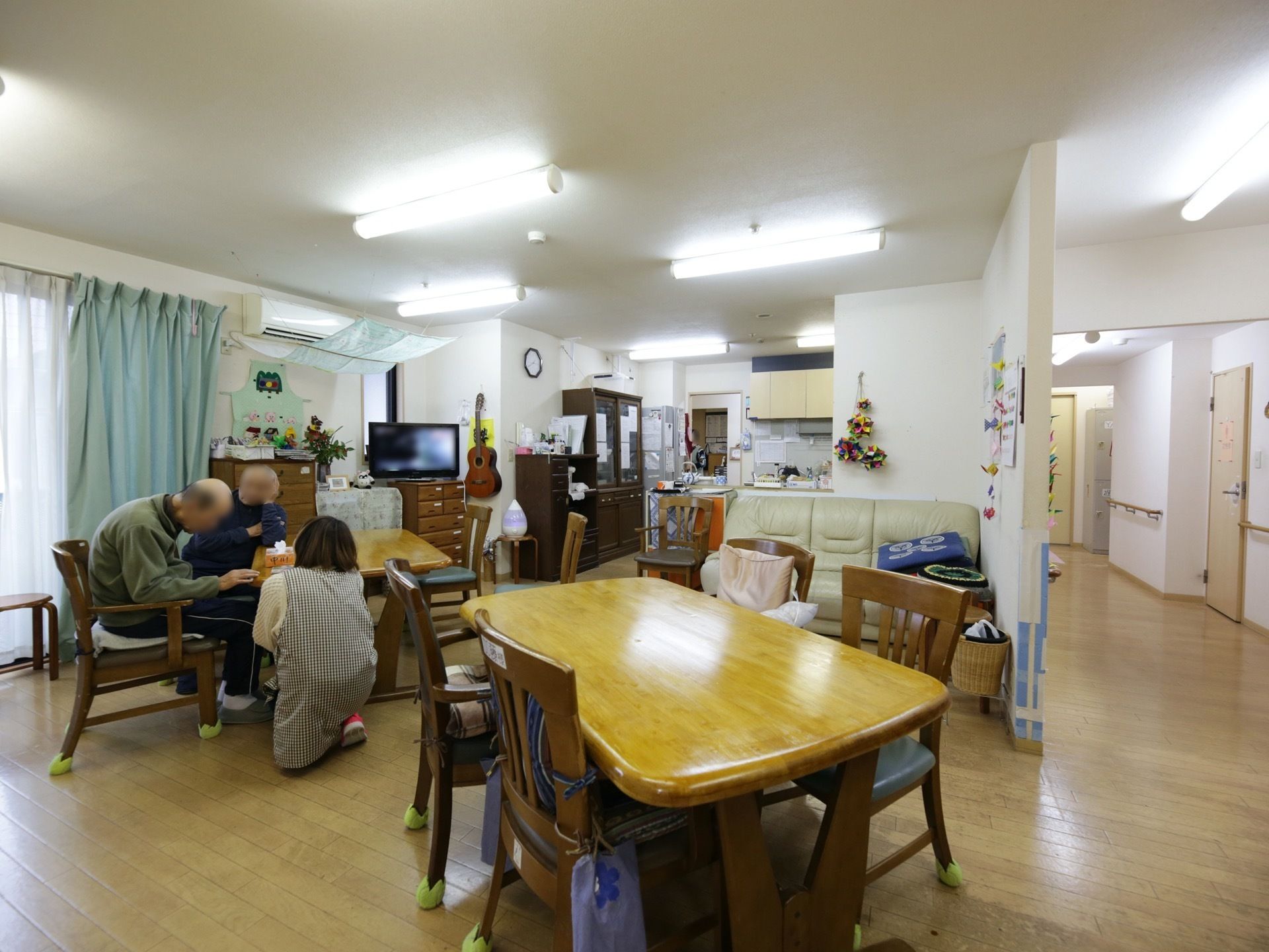 食堂の窓際に置かれたテーブルにある椅子に男性の入居者様が２名座っている。そのそばで女性スタッフがお世話をしている。