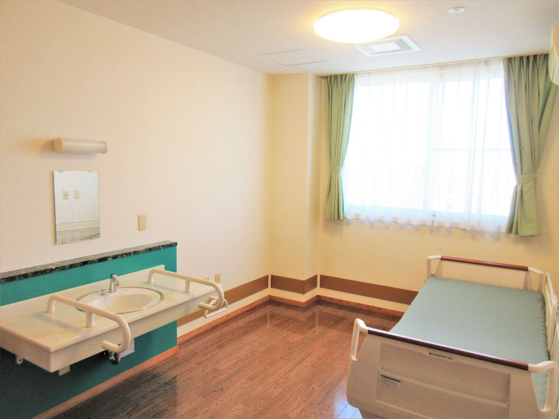個別の部屋にベッドと洗面台がある様子。窓がありカーテンがかかっているこじんまりとした部屋