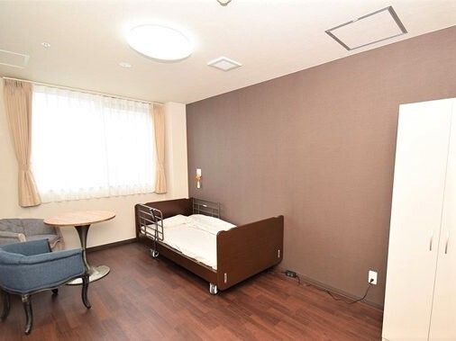 カーテン付きの大きな窓があり、クローゼットやベッド、テーブルセットが置かれ、緊急通報装置も付いている明るい部屋。