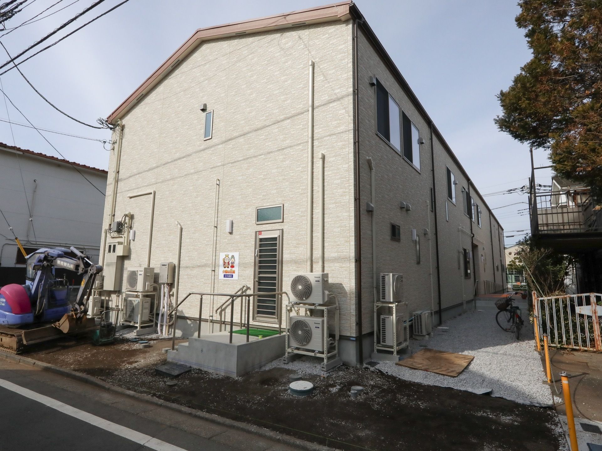 施設は、鉄骨造りの２階建てである。入口に手すり付きの階段がある。建物の脇には空調機の室外機が数台置かれている。