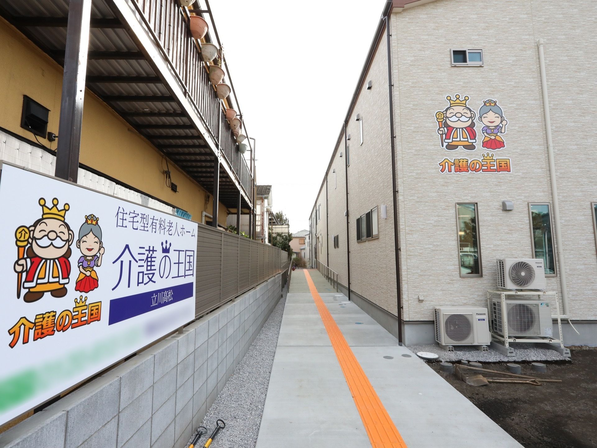 隣との境界ブロック塀に施設名の大きな看板が取り付けてある。施設の建物に沿って、中央に点字ブロックを入れた通路が伸びている。