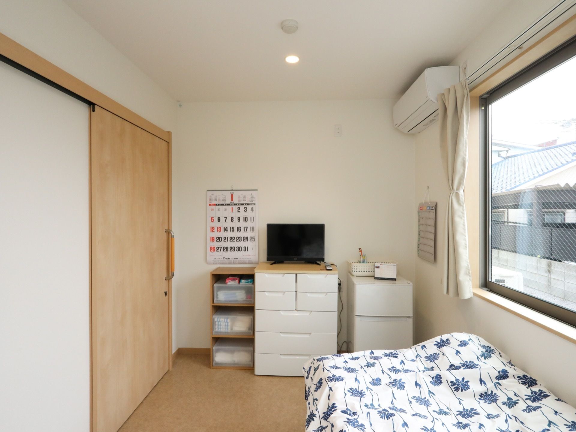 居室の入口ドアはスライド式となっている。広い高窓があり、カーテンが掛かっている。ベッドが置かれ、壁際には整理棚などの家具が並んでいる。