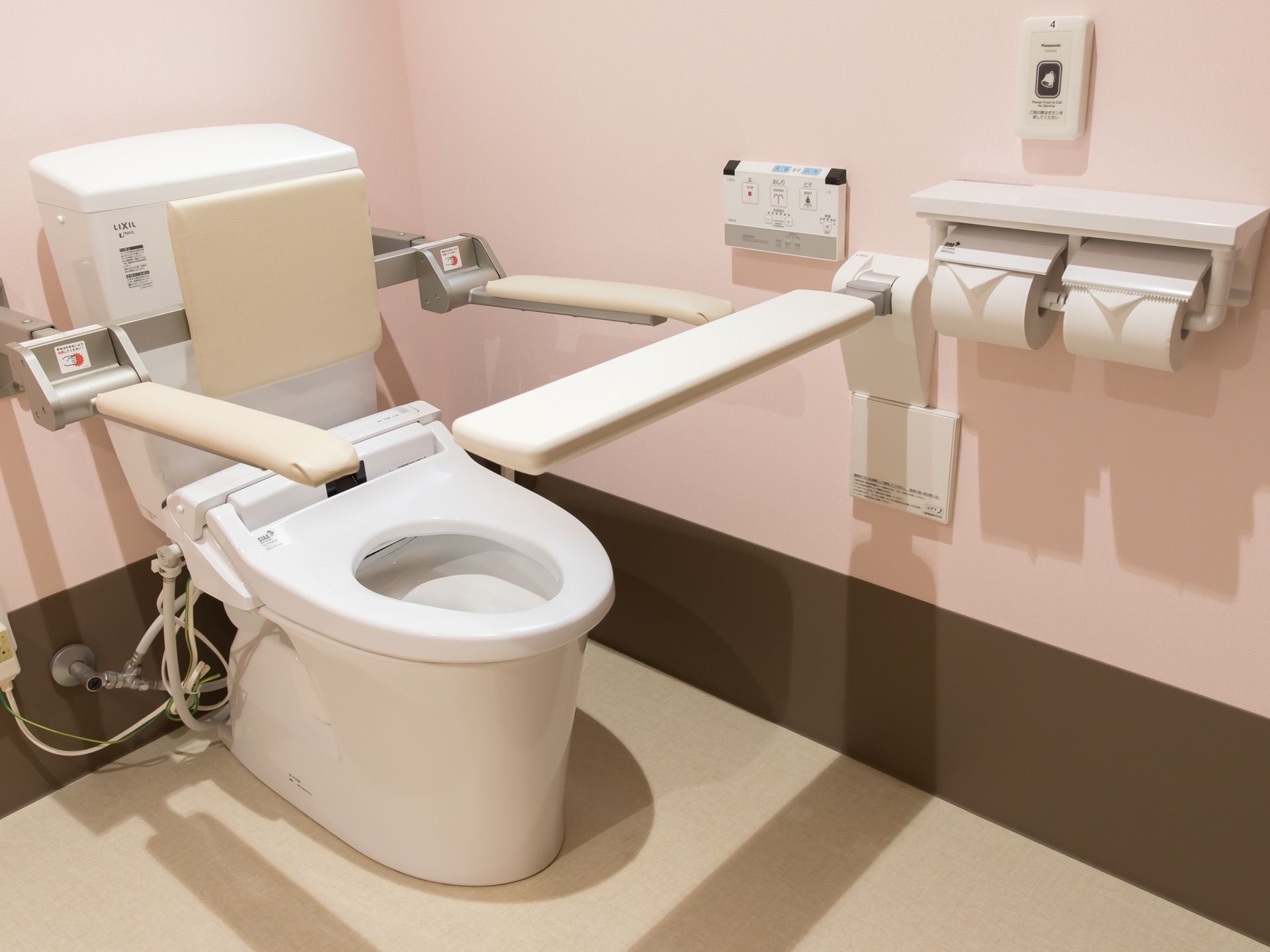 ピンクと茶色の壁に淡い茶色の床のトイレ。背もたれと手すり、温水洗浄便座付きの洋式便器と、緊急呼出しボタンなどが備え付けされている。