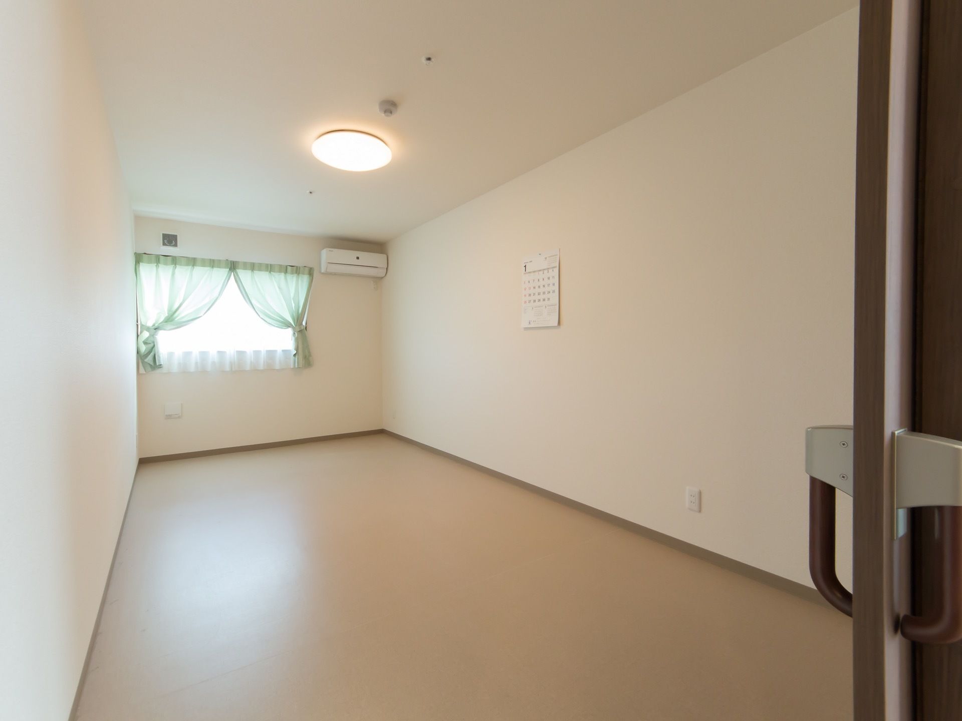 長方形の広い居室。内装は洋風で、白い壁にブラウンの床となっている。壁には窓が1つあり、エアコンががある。