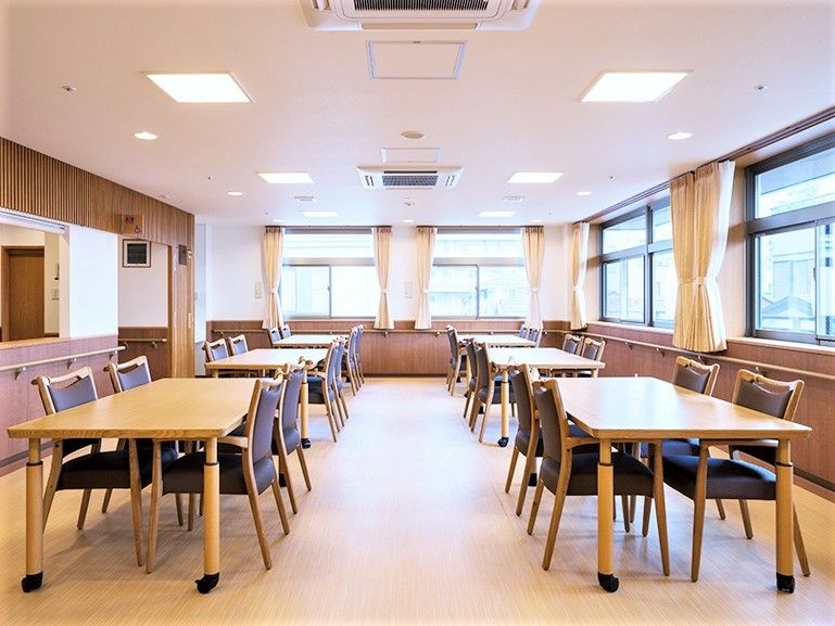 4人掛けのダイニングテーブルがたくさん並び、壁には手すりが設置され、カーテンが付いた大きな窓がある。
