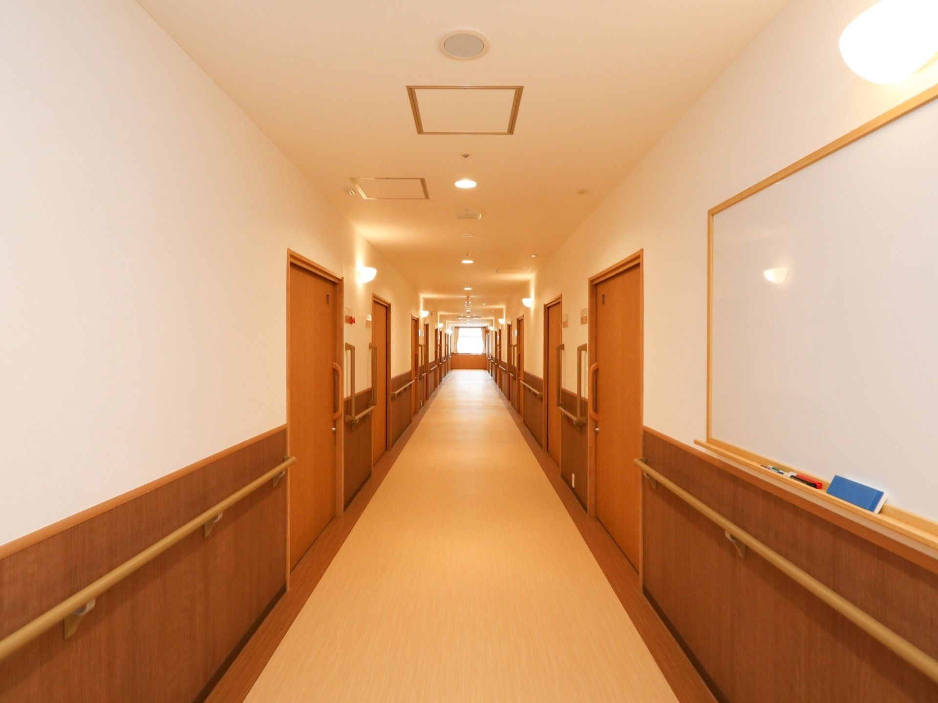 両サイドの壁に手すりが設置されている廊下で、手前にはホワイトボードが掛けられている。天井には等間隔で照明が付けられている