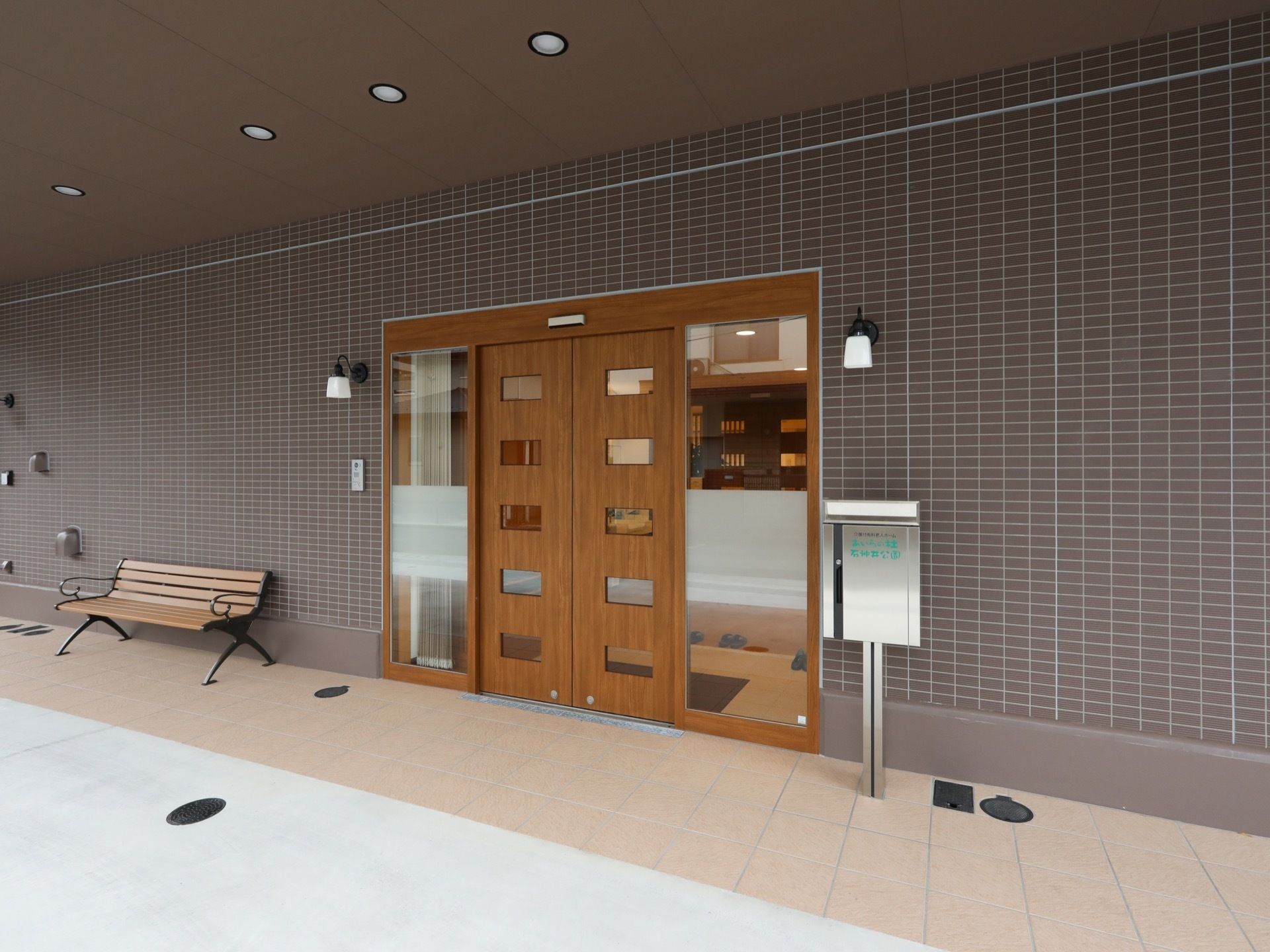 木のぬくもりが感じられる自動ドアが設置されている、屋根付きの玄関。横には、ベンチや郵便ポストが置かれている。
