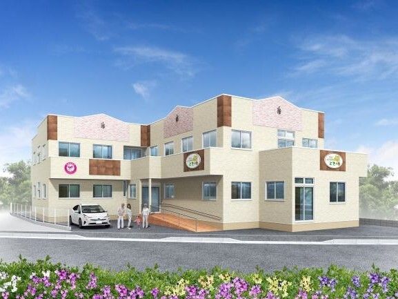ベージュ系の2階建ての建物。玄関には手すり付きのスロープがあり、そのすぐ横に車が止まっている。