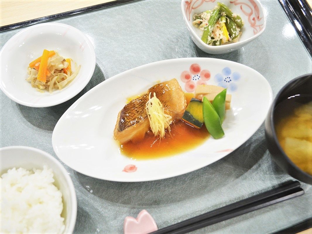 トレーに並べられた食事。煮魚には野菜やしょうがが添えられ、みそ汁や白ご飯、野菜の和え物などがある。