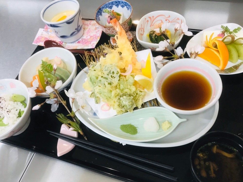 黒いトレーに並べられた食事。豆ご飯に野菜の炊き合わせ、メインの天ぷらにフルーツなど。