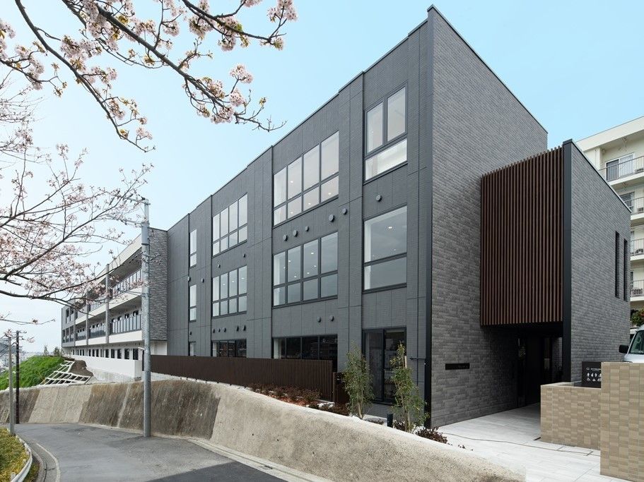 グレーの3階建ての建物の周りには駐車場や木を植えられたスペースがあり、自然も多い。